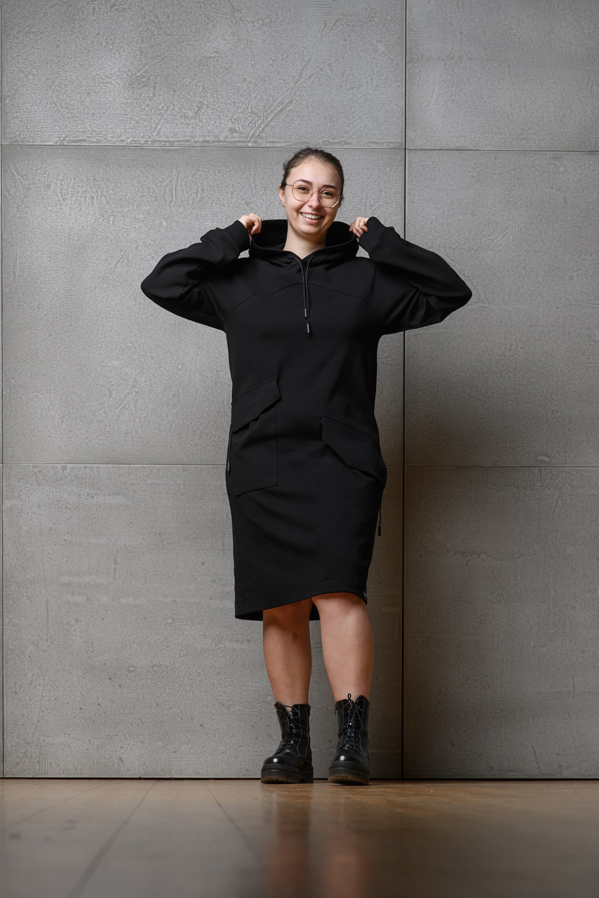 Eine Frau trägt ein schwarzes Hoodiekleid, das bis zum Schienbein geht. Sie lacht in die Kamera und hat die Hände an der Kapuze des Kleides und hebt diese leicht hoch.