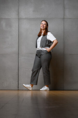 Eine Frau in einem Denim Outfit bestehend aus einer Jeansweste und einer Hose mit Bundfalten aus dem gleichen Denimstoff. Sie hat die Hände in der Hüfte und lacht in die Kamera.