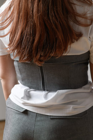 Die Nahaufnahme der Rückenansicht einer Korsage grobem Reißverschluss. Sie ist kurz geschnitten und geht somit nur in die Taille. Darunter trägt das Model ein weißes T-Shirt.