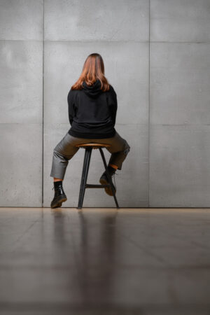 Eine Frau trägt einen schwarzen Oversized Hoodie. Sie sitzt dabei auf einem alten Hocker. Der Rücken ist zur Kamera gewannt. Sie trägt auch eine Jeans mit Bundfalten und aufgenähte Taschen.