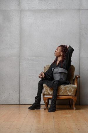 Eine Frau trägt eine Korsage aus Jeansstoff, darunter ein Kleid aus Tencel. Sie sitzt in einem Retro Sessel und schaut nach links.