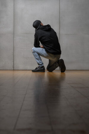 Eine Mann trägt einen schwarzen Oversized Hoodie. Er kniet auf dem Boden und dreht seinen Rücken zur Kamera. Er trägt zusätzlich Blue Jeans und eine Kappe.