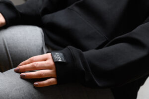 Eine Detailaufnahme des Unisex Hoodies. Die Hand einer Frau ruht auf dem Oberschenkel und zeigt dabei das Etikett am Ärmelsaum.