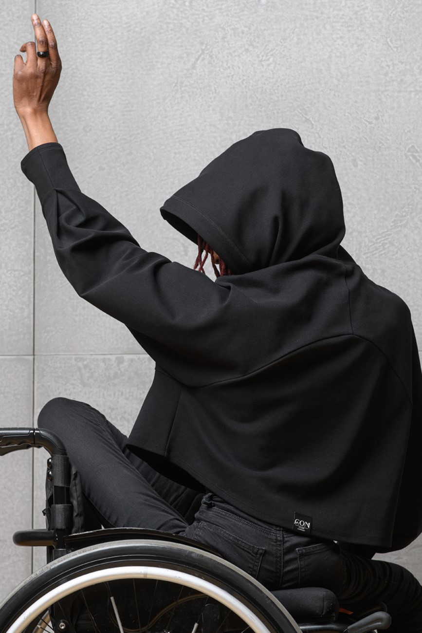 Eine Frau im Rollstuhl trägt einen schwarzen kurzen Hoodie. Sie sitzt mit dem Rücken zur Kamera und hat die Kapuze auf dem Kopf. Dabei hebt sie einen Arm.