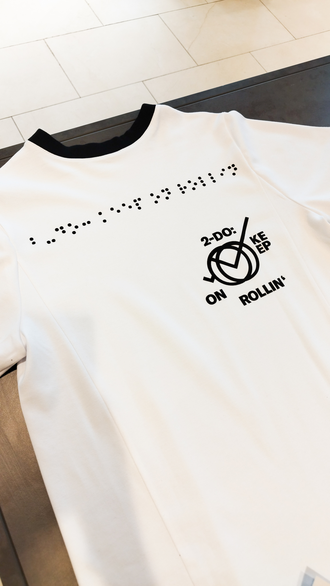 weißes T-Shirt mit schwarzem Druck. Der Druck zeigt ein Design mit dem Spruch: 2-Do Keep on rollin'. Dieser Druck ist in Brailleschrift übersetzt und ist groß auf das T-Shirt gedruckt.