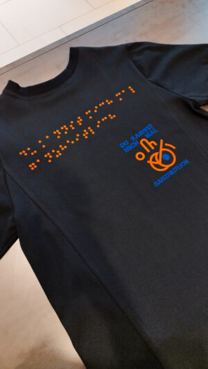 schwarzes T-Shirt mit orange-blauem Druck. Der Druck zeigt ein Design mit dem Spruch: Du kannst mich mal ganzheitlich. Dieser Druck ist in Brailleschrift übersetzt und ist groß auf das T-Shirt gedruckt.