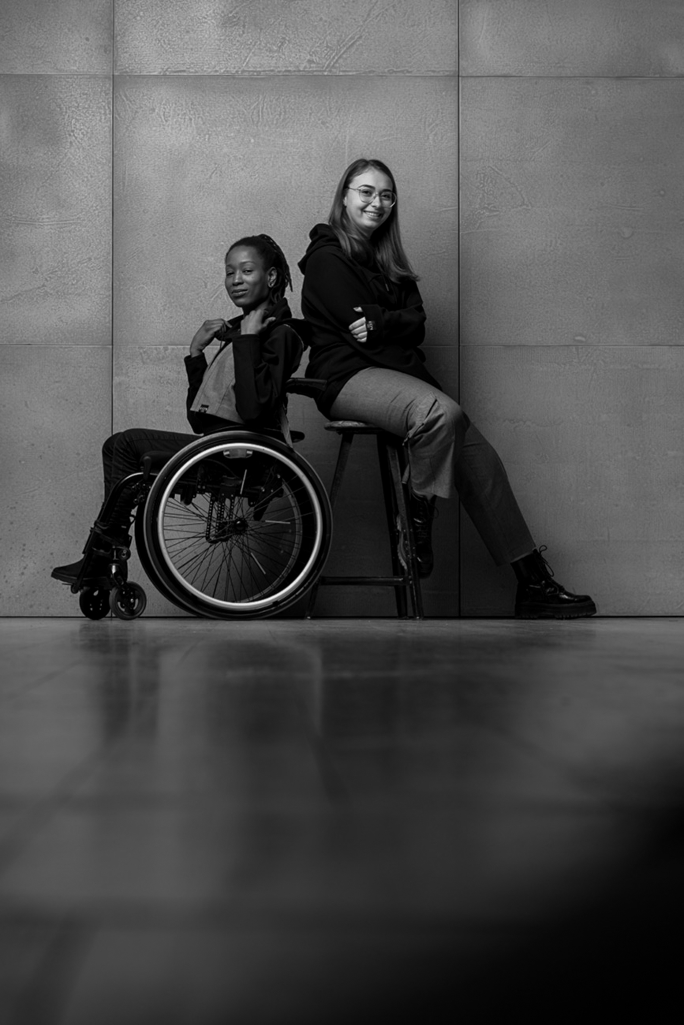 zwei Personen sitzen nebeneinander. Die linke Frau ist schwarz und sitzt im Rollstuhl. Die Frau af der rechten Seite ist rothaarig und sitzt auf einem Barhocker. Beide tragen die inklusive Kollektion von Claire Common