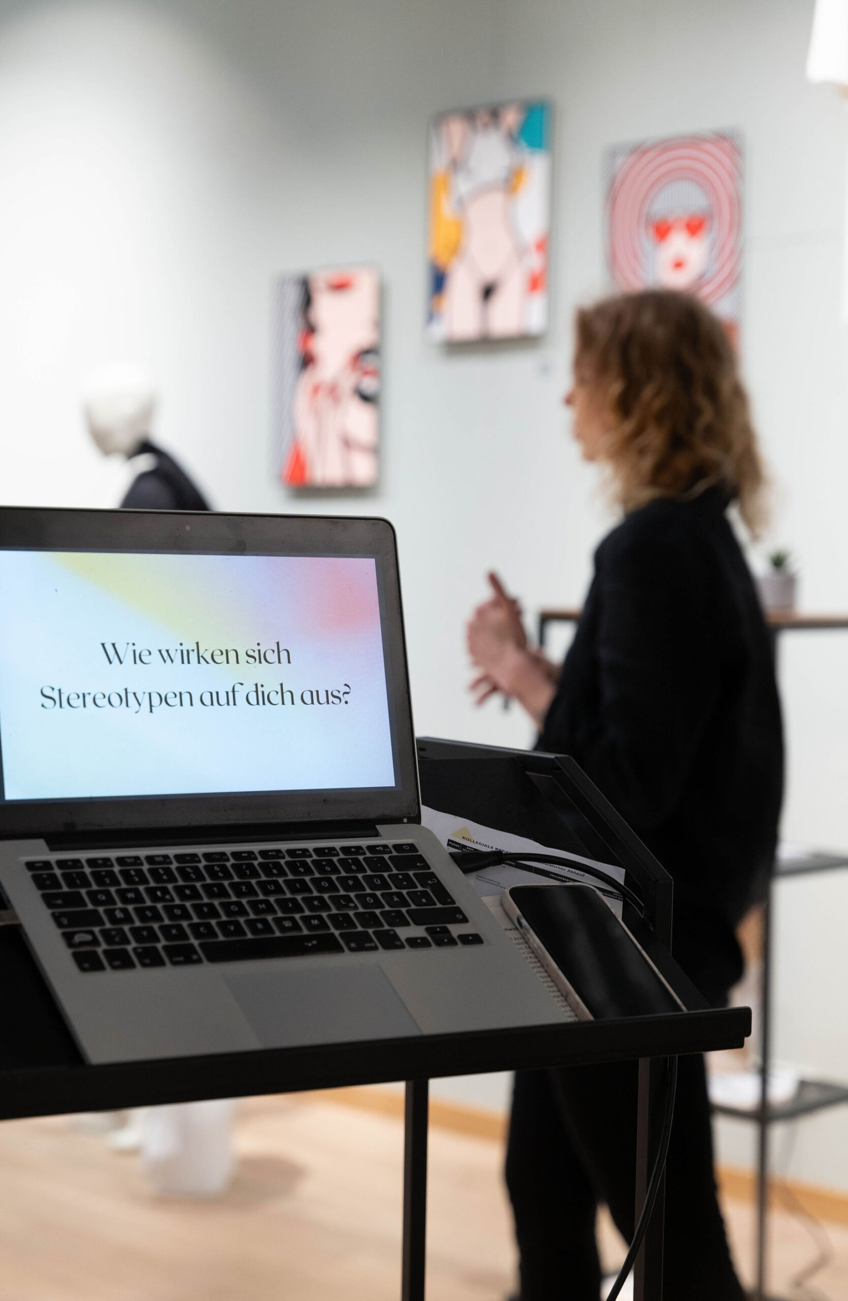 eine Frau mit Locken - Claire Common - steht im Hintergrund und erklärt dem Publikum etwas. Im Vordergrund ist ein Laptop und eine Präsentation zu sehen. Auf der Präsentation steht: Wie wirken sich Stereotypen auf dich aus?