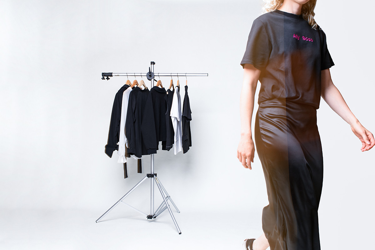 eine blonde Frau mit Locken und schwarzem Outfit läuft durch das Bild. Hinter ihr ist eine Kleiderstange mit Bekleidung