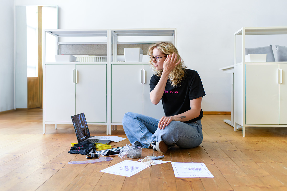eine blonde Frau mit Locken - Claire Common - sitzt auf dem Boden und arbeitet. Vr ihr liegen ein Laptop sowie Blätter, Stoff und Scheren sowie Nadeln.